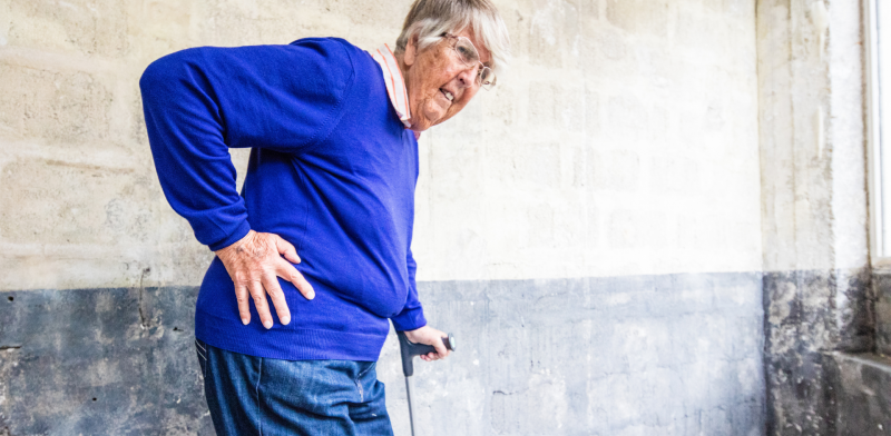 Mujer mayor camina con muleta mientras se lleva una mano a la cadera